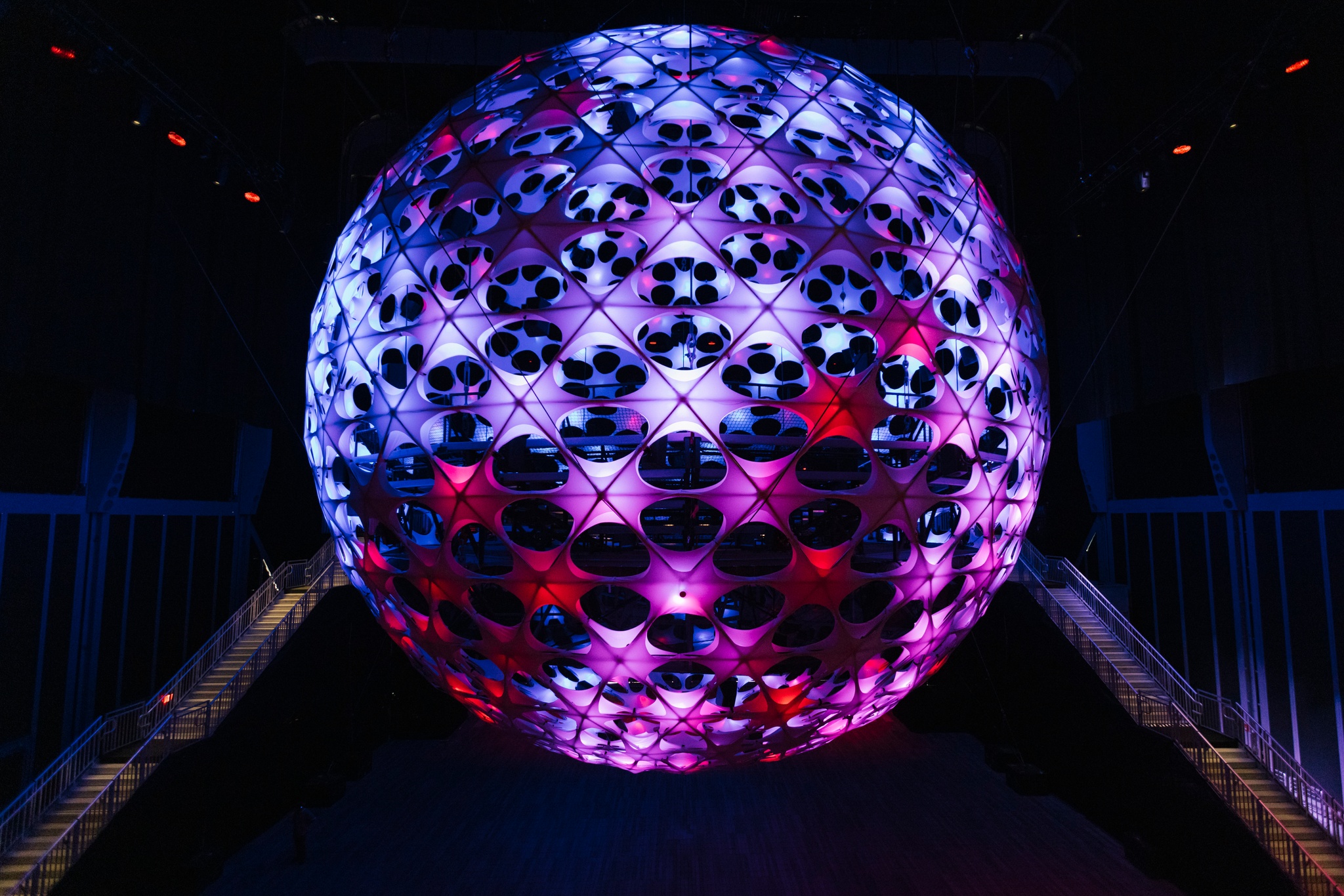 A vast spherical concert hall hangs from a 115-foot ceiling in a dark performance space. The sphere is dotted with nodes that light up in purple and blue light. Flanking the sphere are two sets of metal staircases that rise to the equator of the sphere.