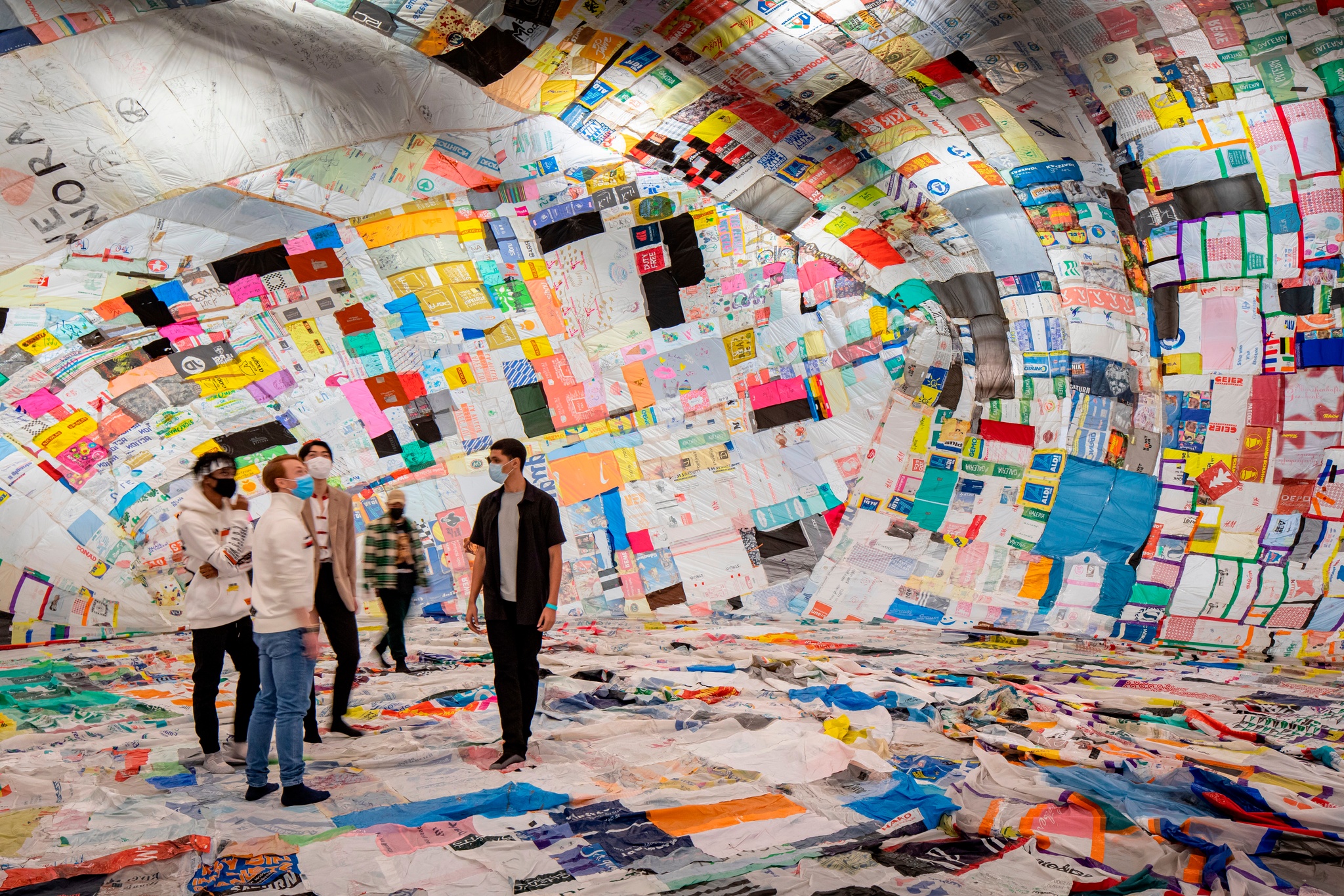 Five people stand inside a cavernous ballon made of a colorful patchwork of reused plastic bags quilted together with tape