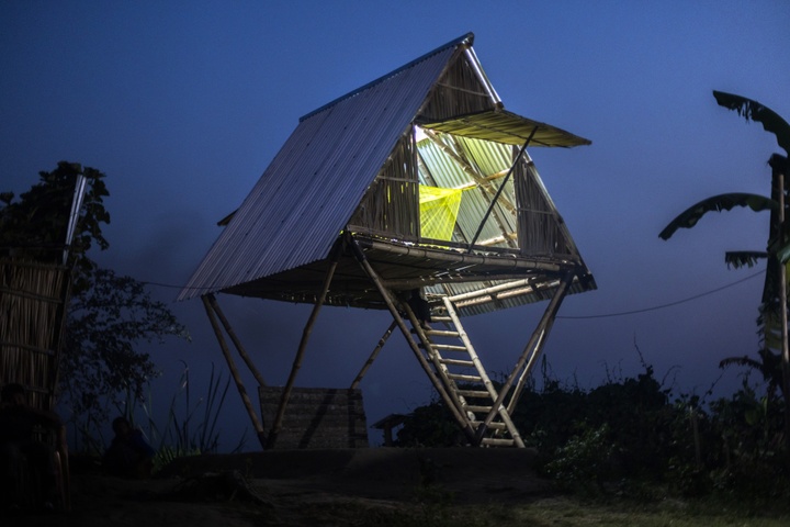Photo showing a triangular structure built from wooden poles, brightly illuminated from the inside at dusk.