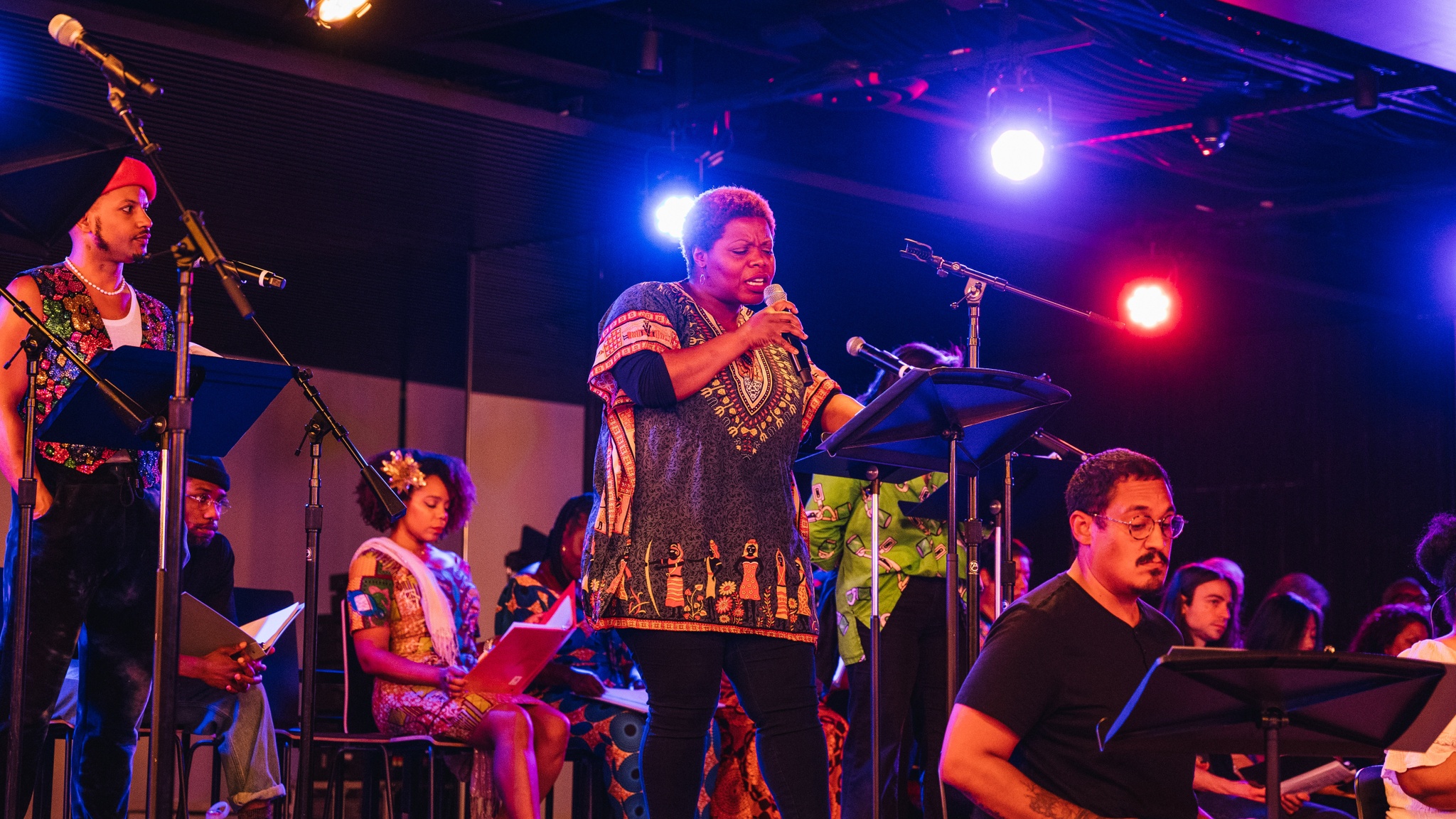 A Black woman with short hair stands on a stage holding a microphone that she speaks into. She looks down at a script she is reading from. Around her are other participants in the reading, and she is lit by blue stage lights in the ceiling behind her.