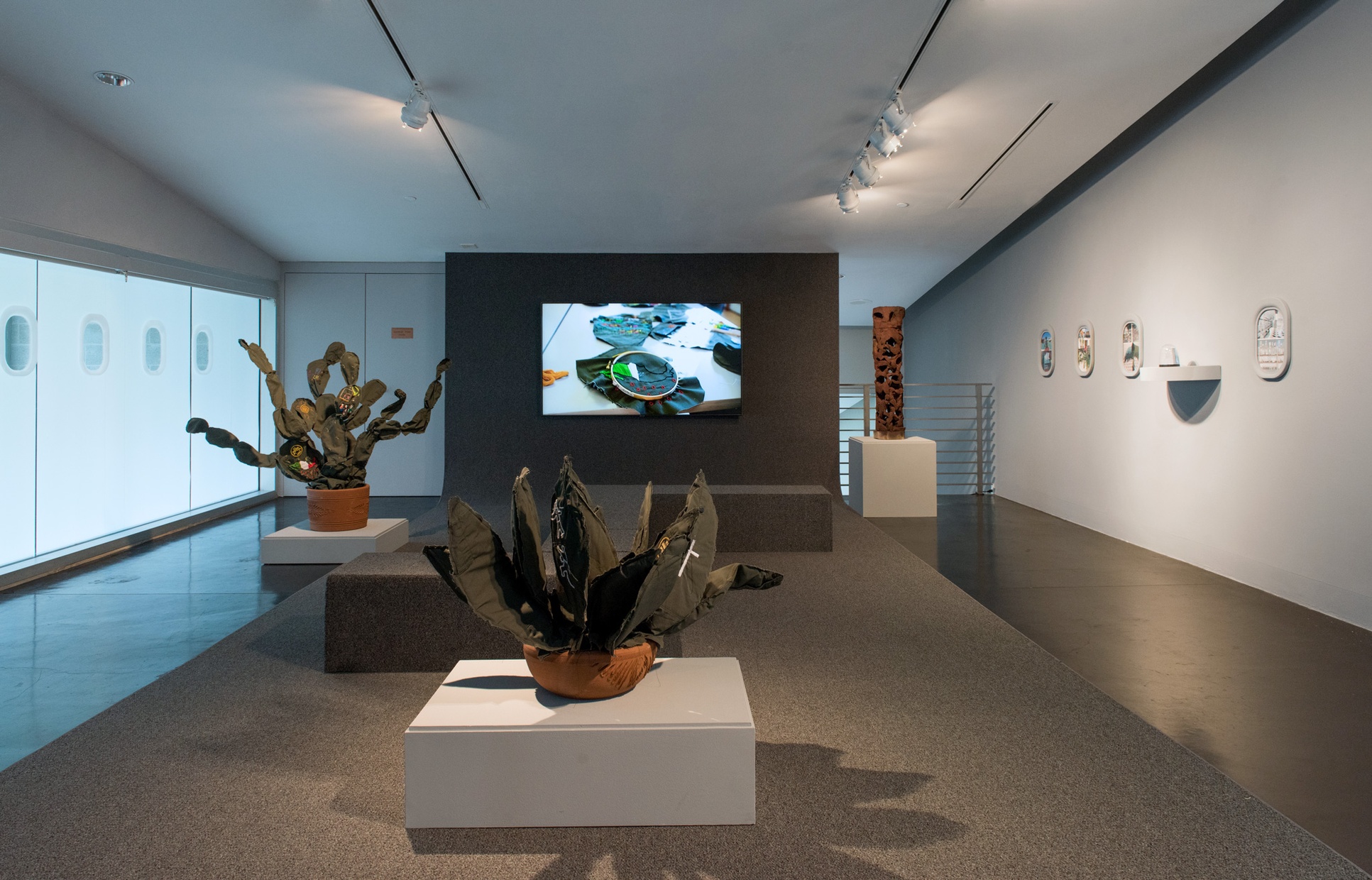 A room with a wall of windows on the left, soft cactus sculptures in the center with a television monitor playing video on the wall behind them, and art hanging on the wall on the right.