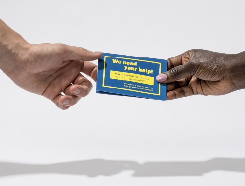 A hand handing a blue card with yellow text 'We Need Your Help' on to another hand over a plain white background
