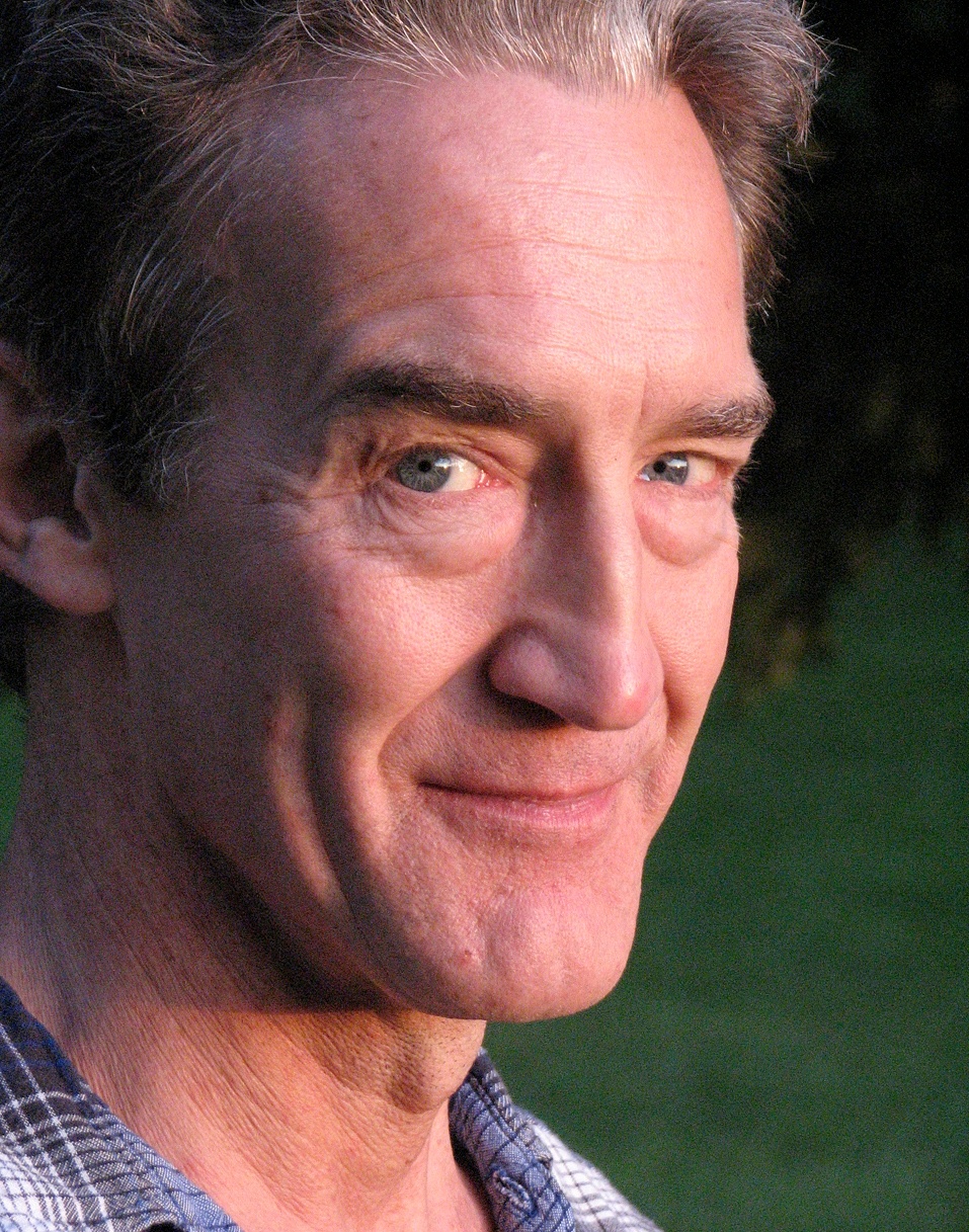 A portrait of actor Guy Paul, a white man seen in three quarter profile smirking at the camera
