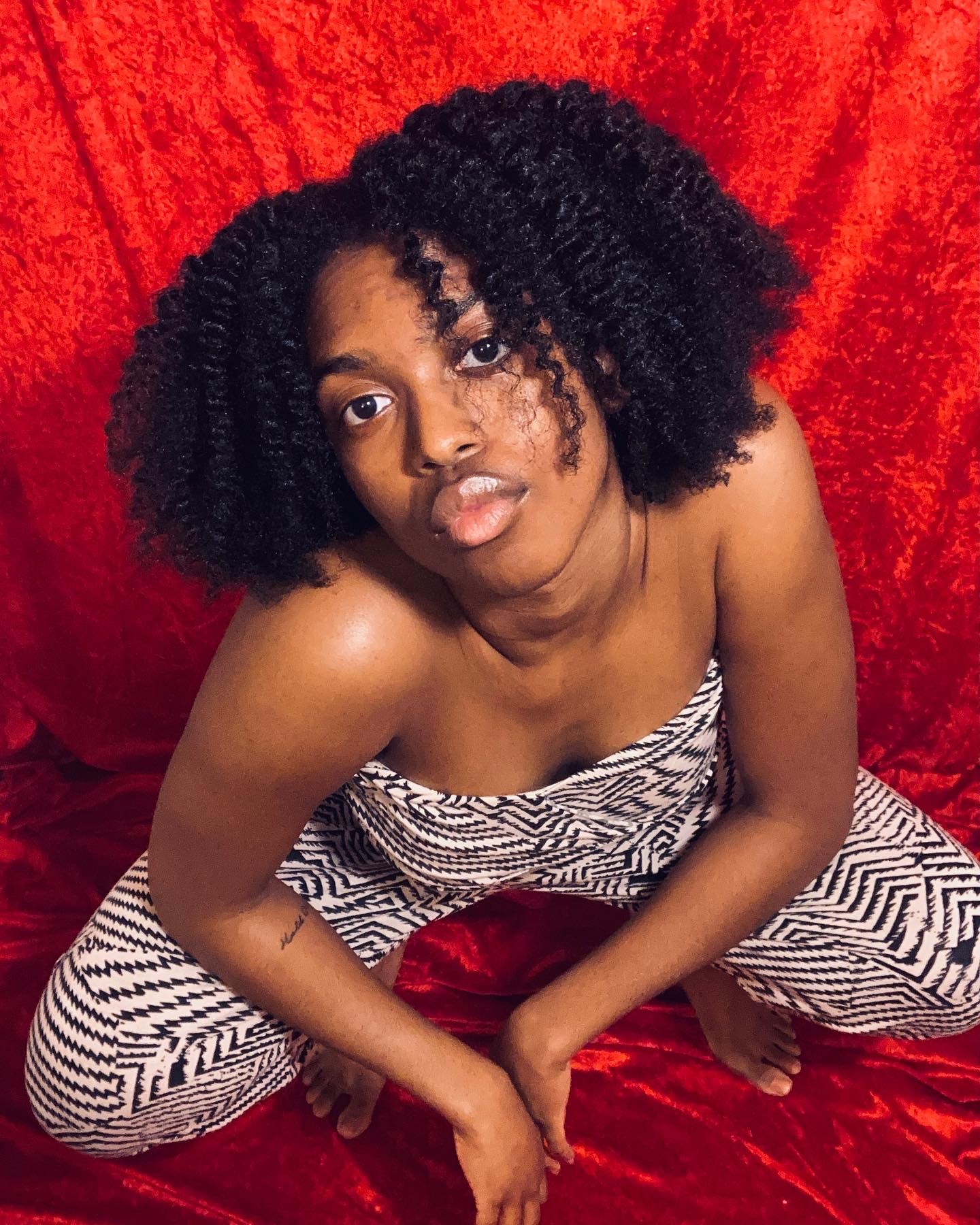 A Black woman crouching on a red photo studio backdrop and looking up at the camera. She has curly hair parted in the middle and wears a shoulderless jumpsuit with a black-and-white zigzag pattern.