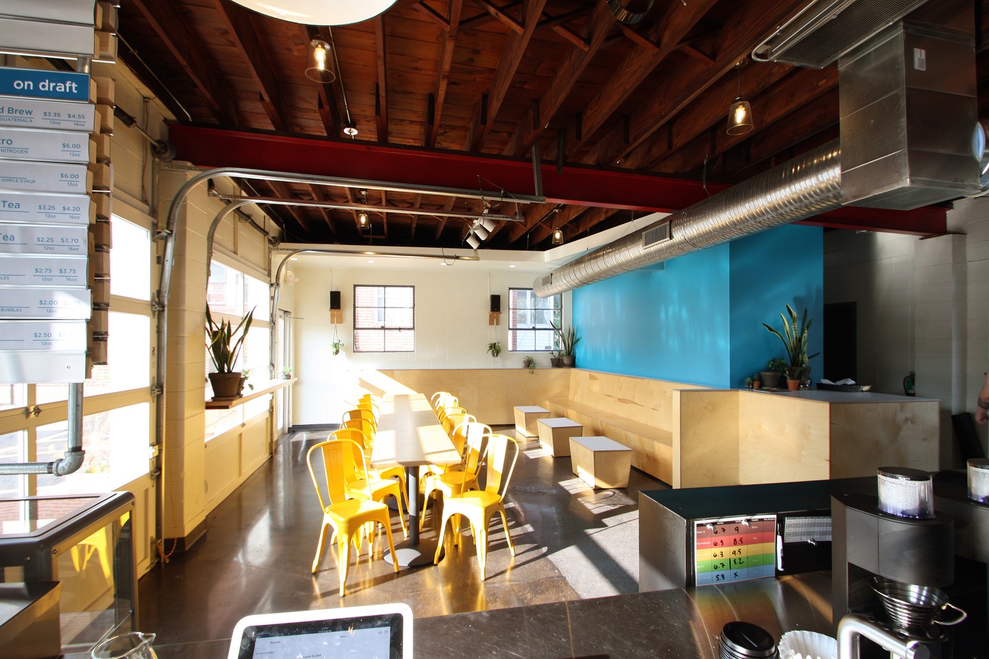 Interior of a coffee shop with exposed wooden beams, garage doors along one wall, pine wood tables and bench seating, and a baby blue accent wall.