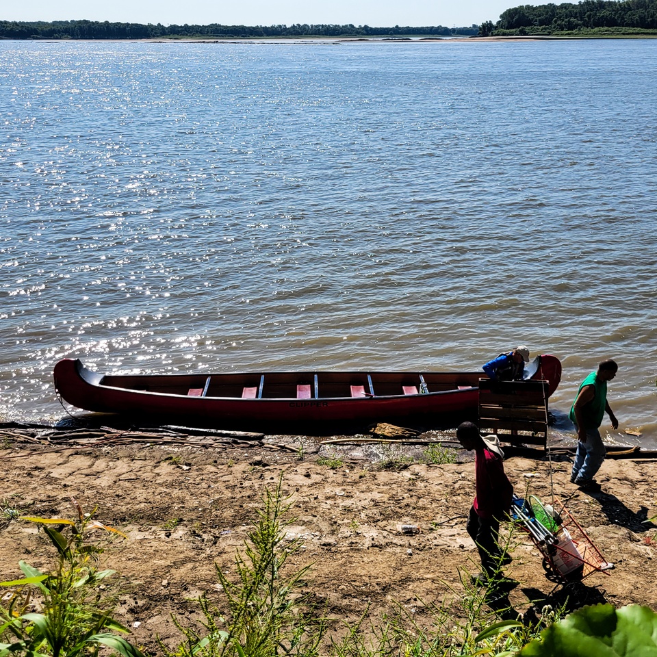 A red canoe by a river bank with 3 figures on the right 