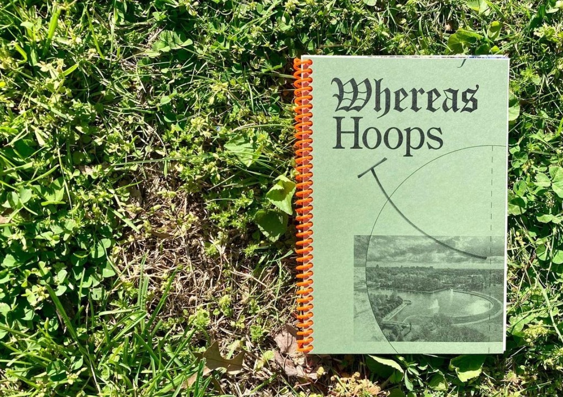A moss green book with red spiral binding titled Whereas Hoops lies on a bed of green grass.