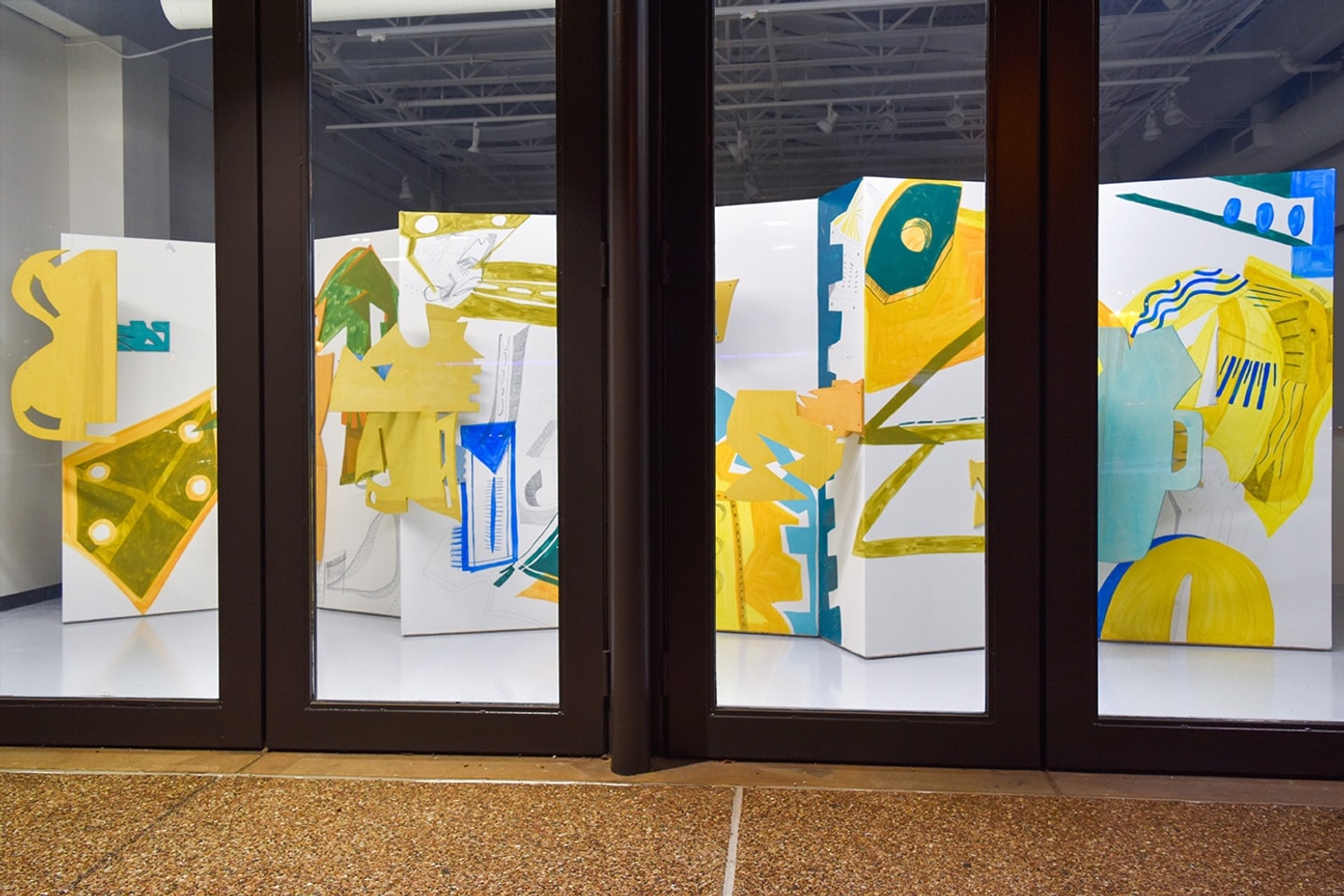 A wide view of an art installation by Ribaudo, seen through large glass gallery doors, featuring multi-paneled white partition walls decorated with yellow, green, and blue abstract cutouts and painted motifs.