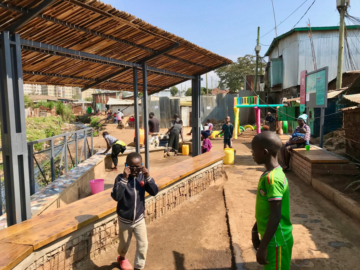 Photo of people, mostly children, in an outdoor area with a long, wooden bench and a shade covering.