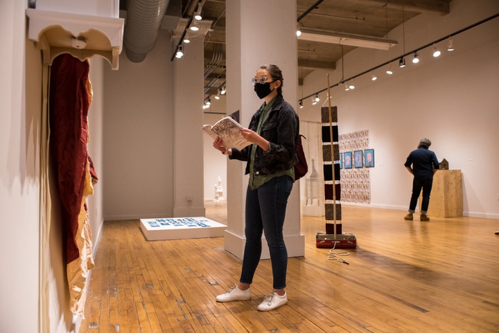 Person in facemask consults an exhibition catalog while looking at an artwork on the wall.