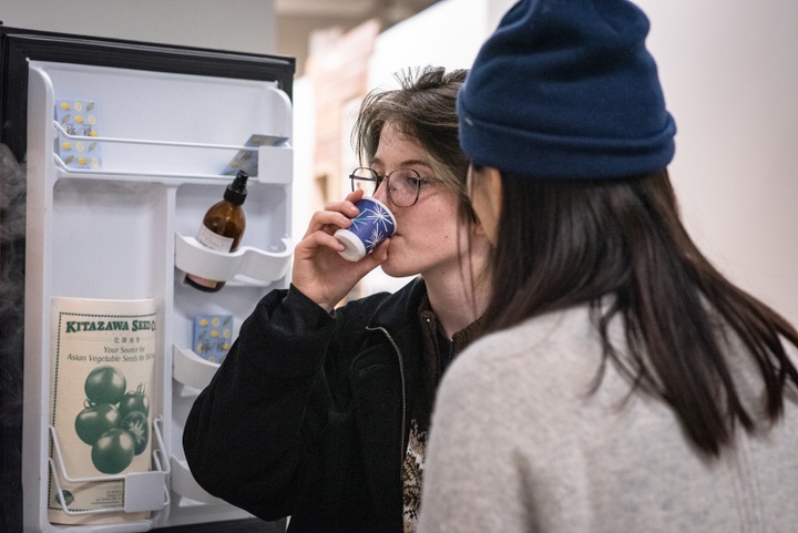 Person sips out of a small paper cup in front of an open fridge with a seed catalog in its door.