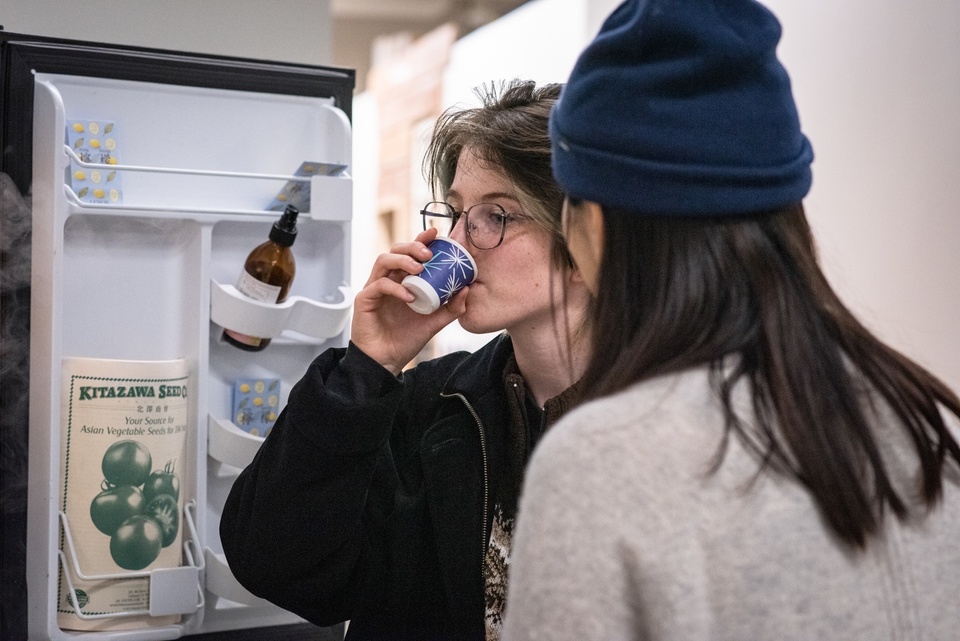 Person sips out of a small paper cup in front of an open fridge with a seed catalog in its door.