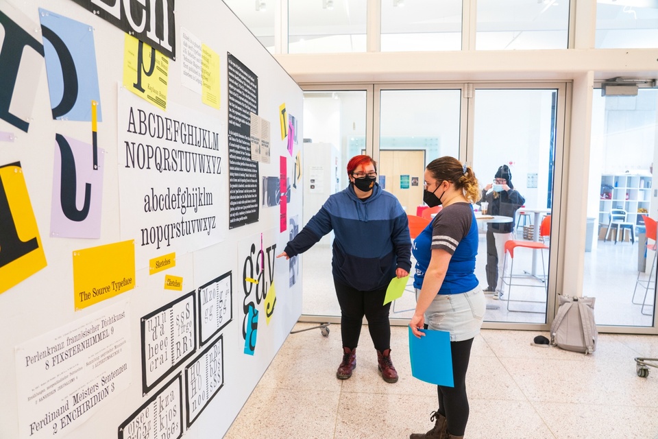 Two students look at a wall of samples of a modern serif font.