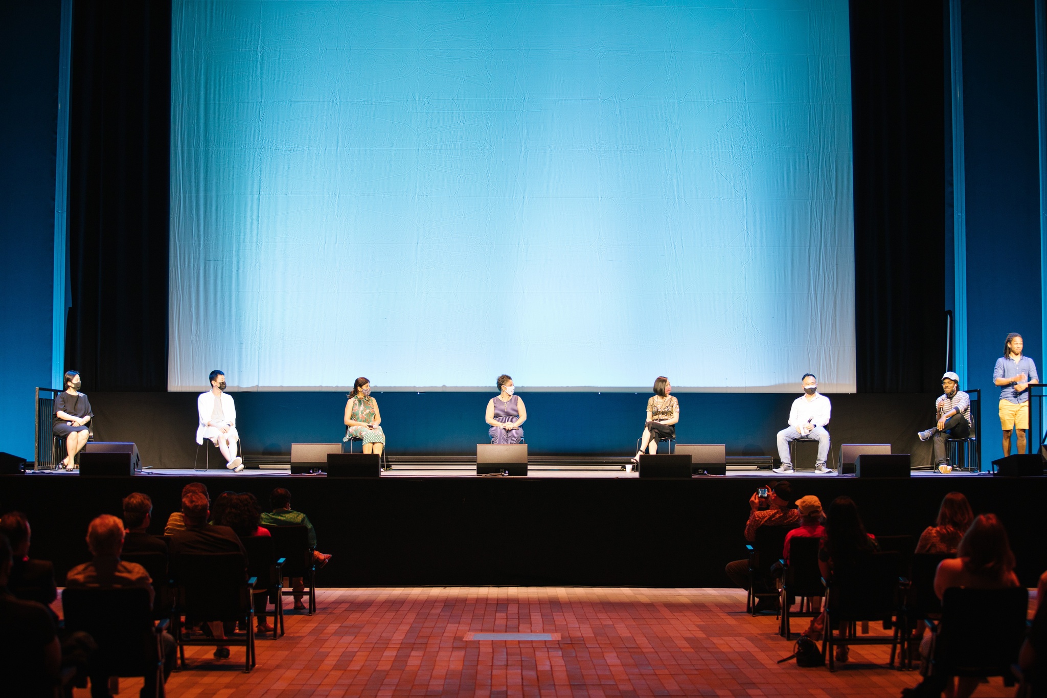 A line of seven panelists sit on a stage facing an audience with a film screen rising high above them in the background. 