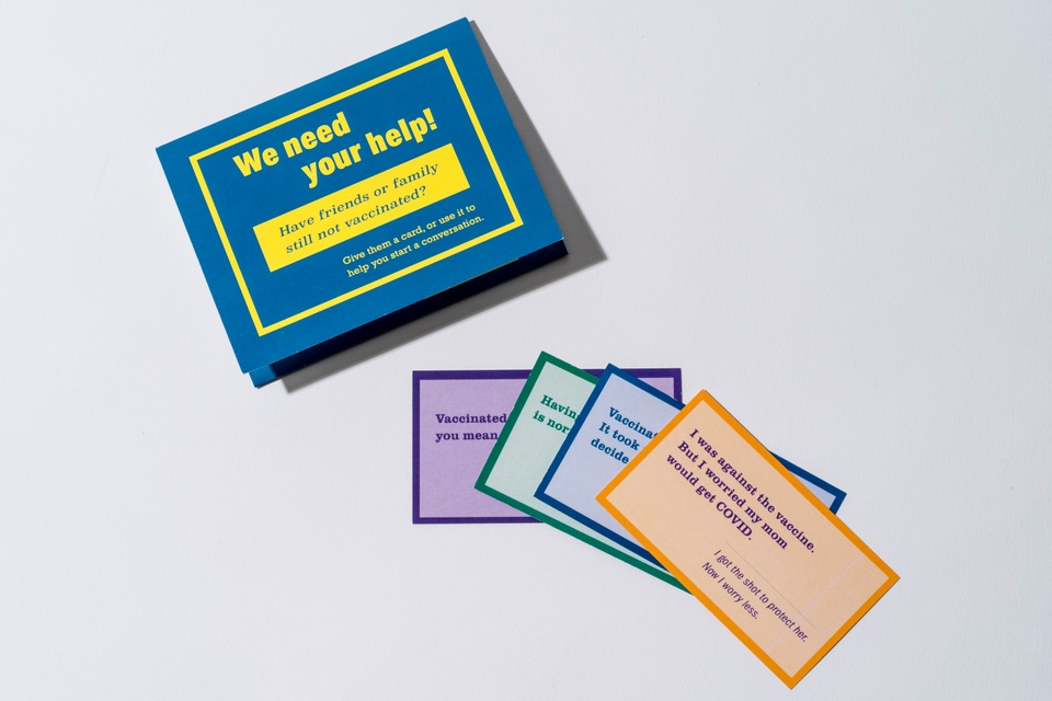 A blue and yellow card holder lies on a white background next to 4 conversation cards splayed out, going in order of purple, green, blue and orange. The card holder reads "We need your help! Have friends or family that are still not vaccinated? Give them a card or use it to start a conversation."