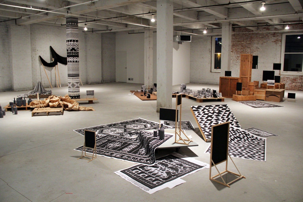 Multiple papers with black and white screenprints scattered on a concrete floor with stacks of wooden boxes and bottles in the background.