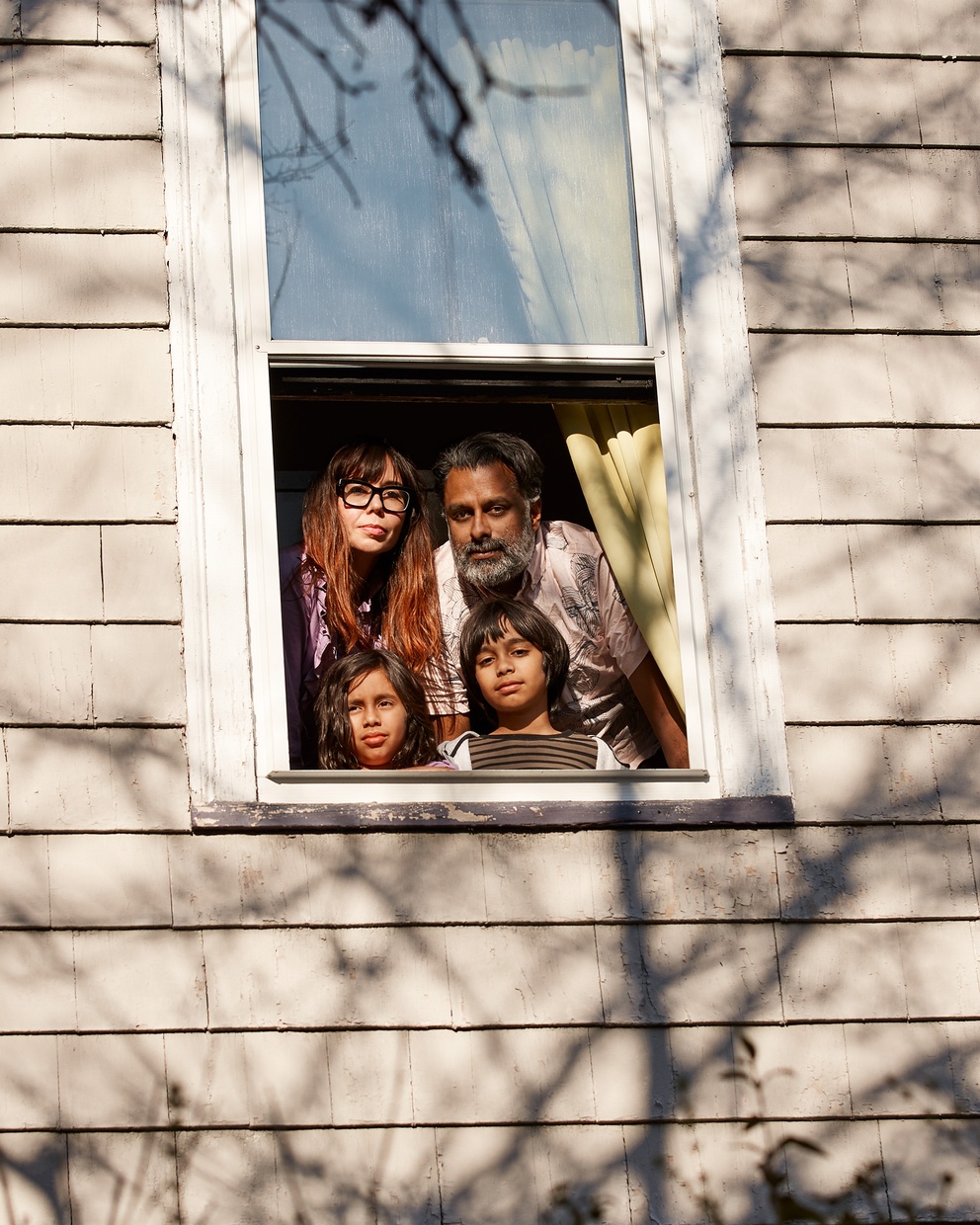 A color photograph of the side of a tan house with an open window and a family of four looking out.