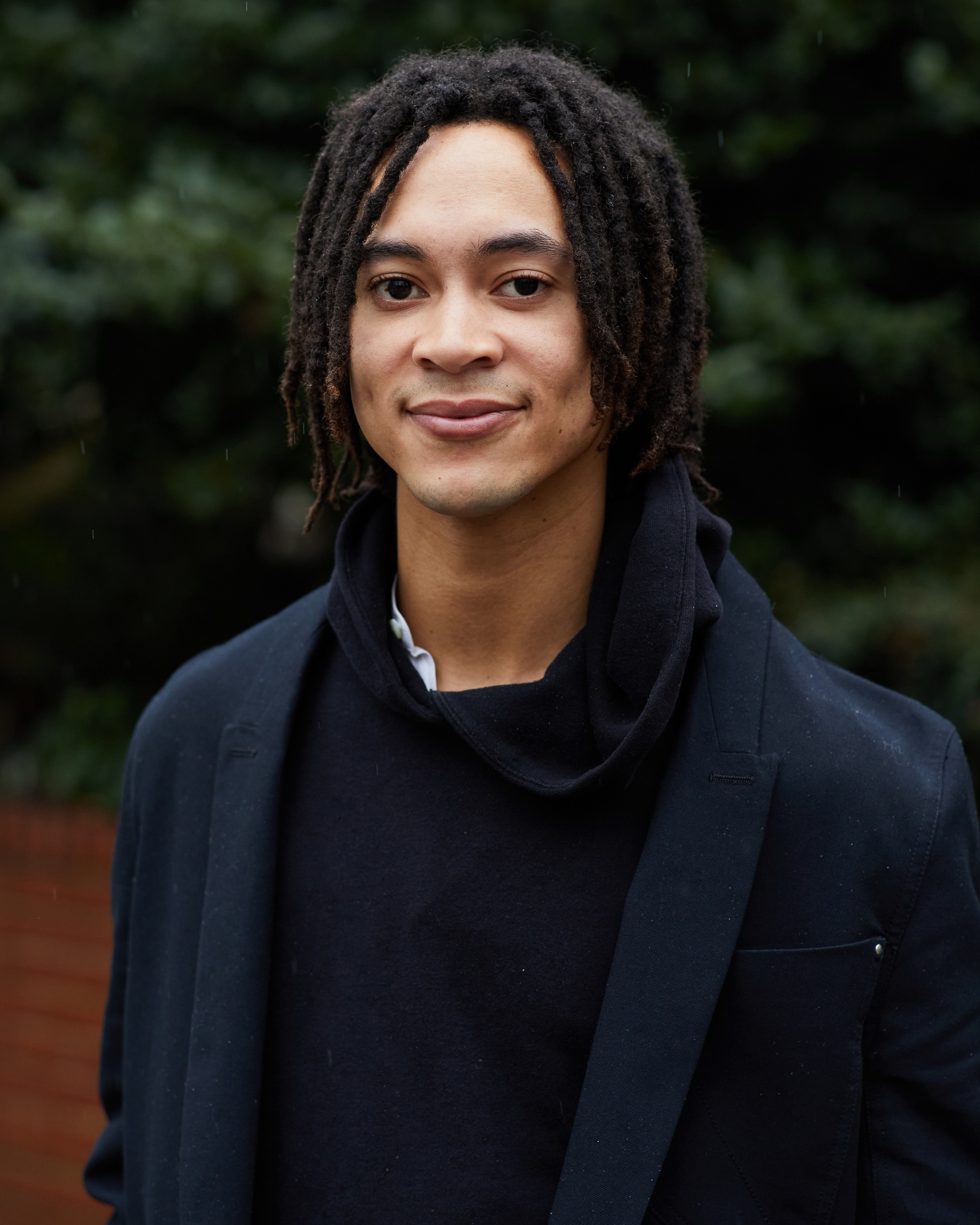 This is a portrait of Sandy Williams IV. They pose with the trees lining a street behind them. They are wearing a dark blazer over a dark hoodie and have their hair in short locs that fall to their jawline.