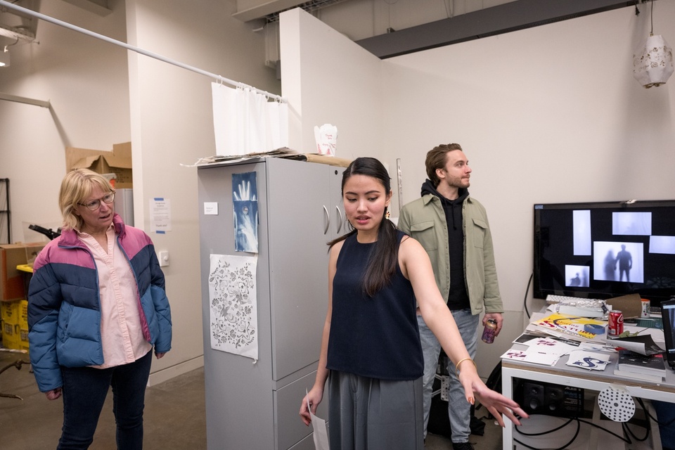 Two visitors enter a student's studio space. The student points something out to them in the bottom corner of the frame. 