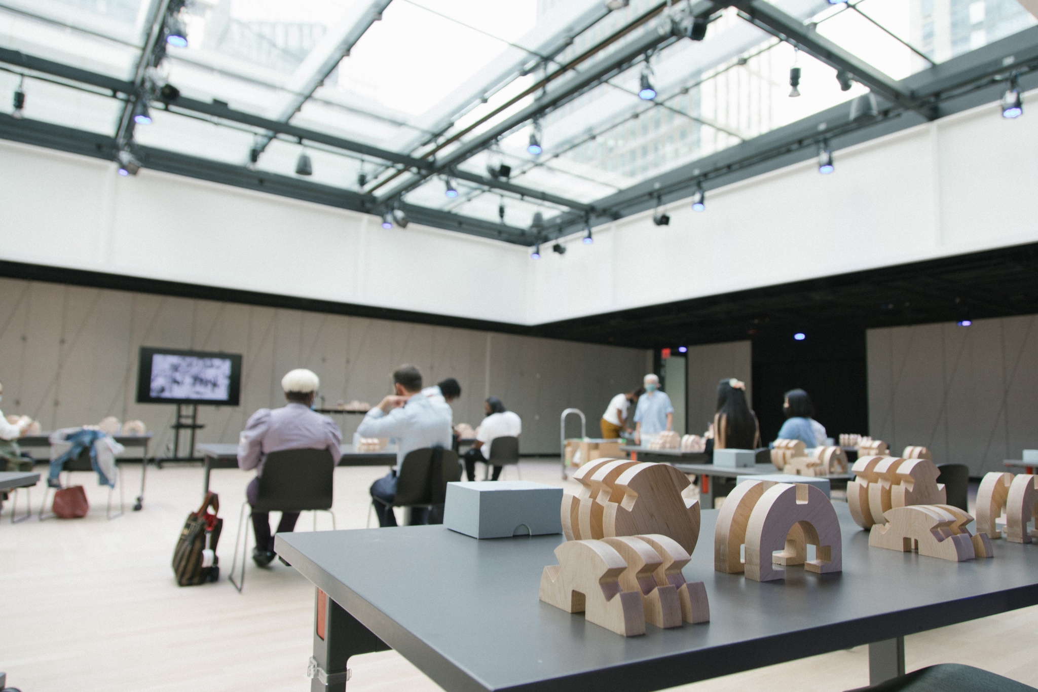 Long tables arranged in rows with circular wooden blocks on them. Some tables have participants seated at them for a workshop centered on these blocks.