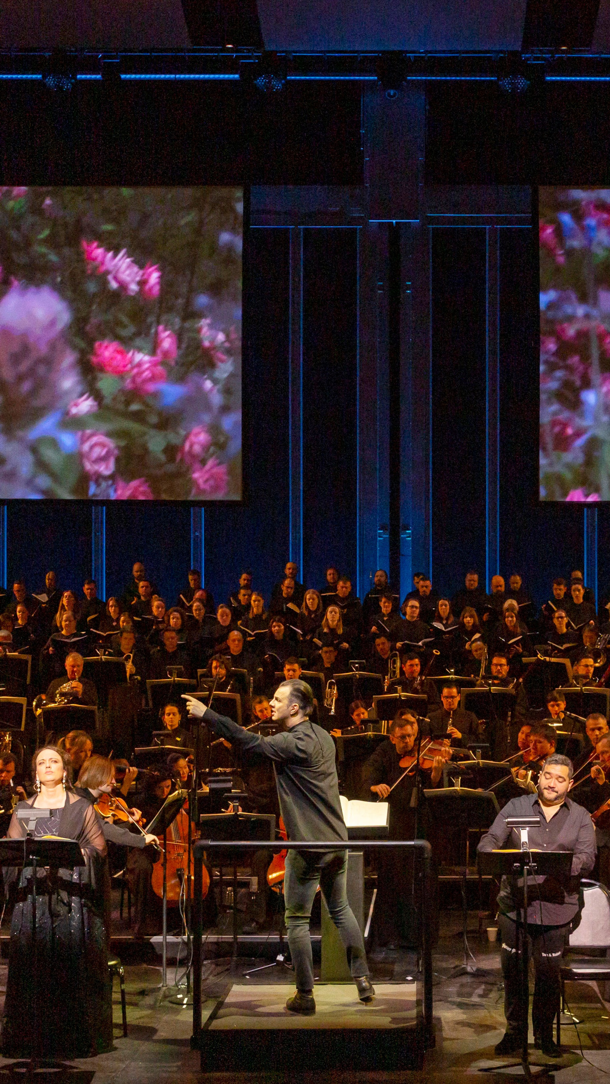 Teodor Currentzis conducting an orchestra in front of two large screens.