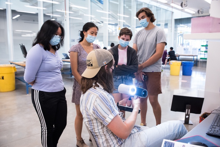 Group of students gathers around a shop tech holding up a glowing handheld 3D scanning tool.