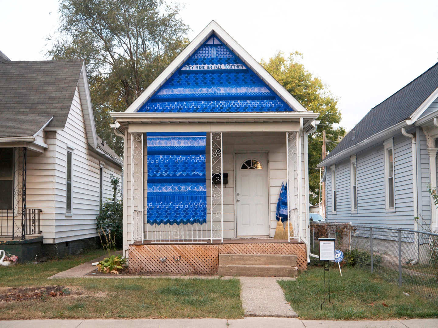 A textile placed on the façade of a house. The textile consists of a blue and white geometric pattern, with the text "THEY RISE OR FALL TOGETHER" — the fabric also hangs on the front of the house above two brooms.