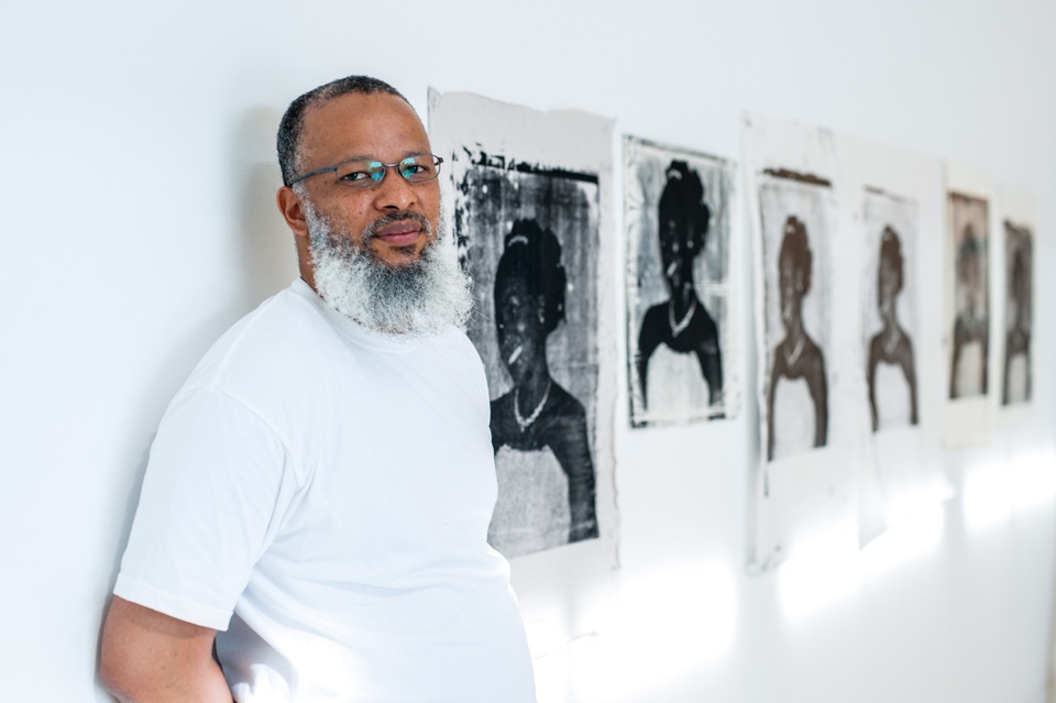 Portrait photo of Meleko Mokgosi in a gallery with framed, black-and-white artworks on white walls.