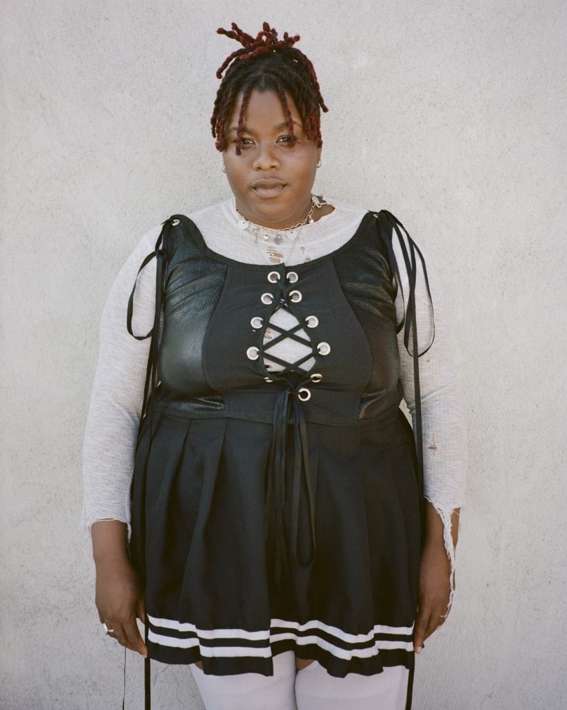 A Black person wearing a short black dress with a lace-up top over a white long-sleeve t-shirt and white leggings. They stand against a black white wall. 
