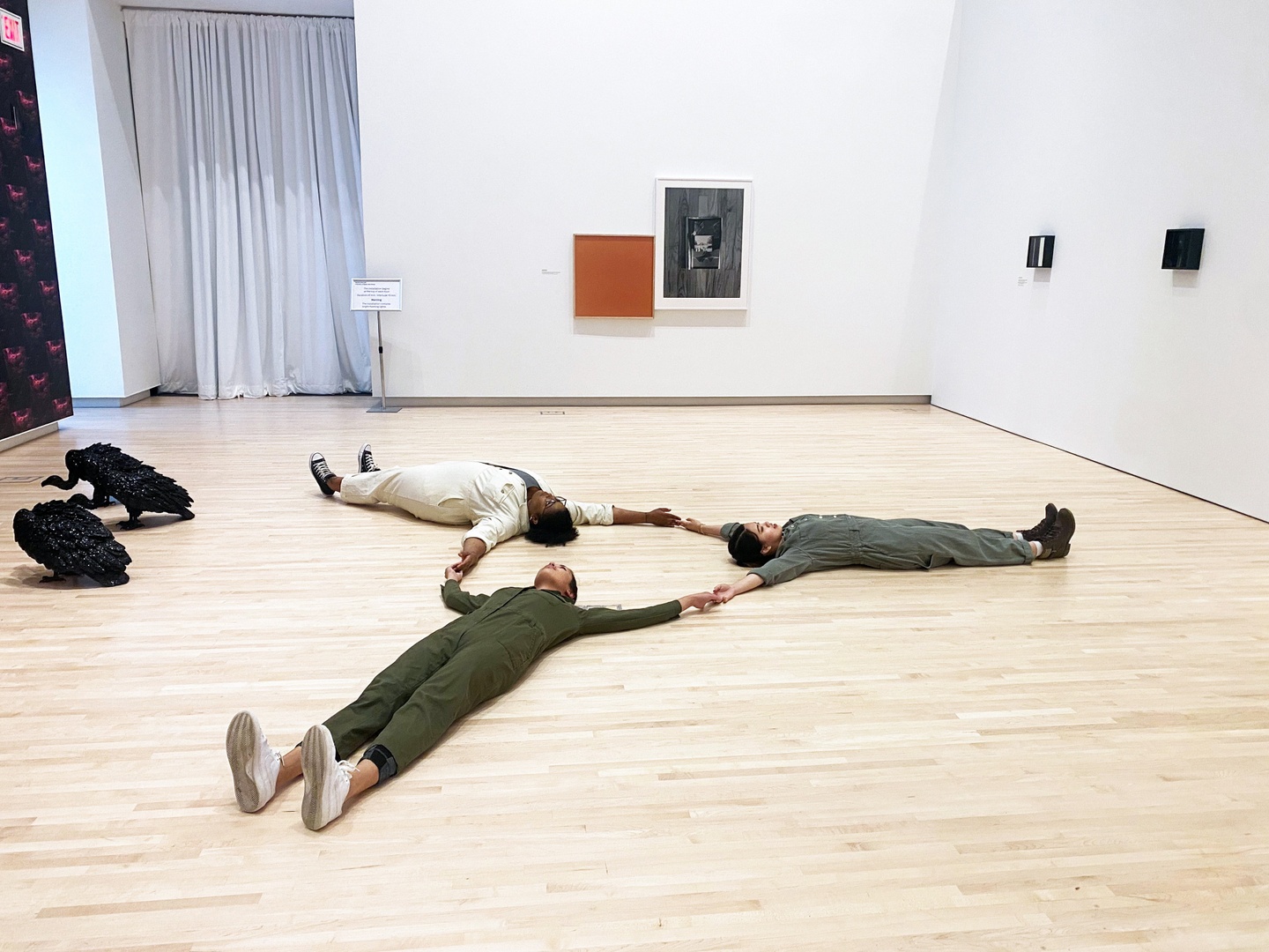 Three students laying on the gallery floor in a triangular formation holding hands