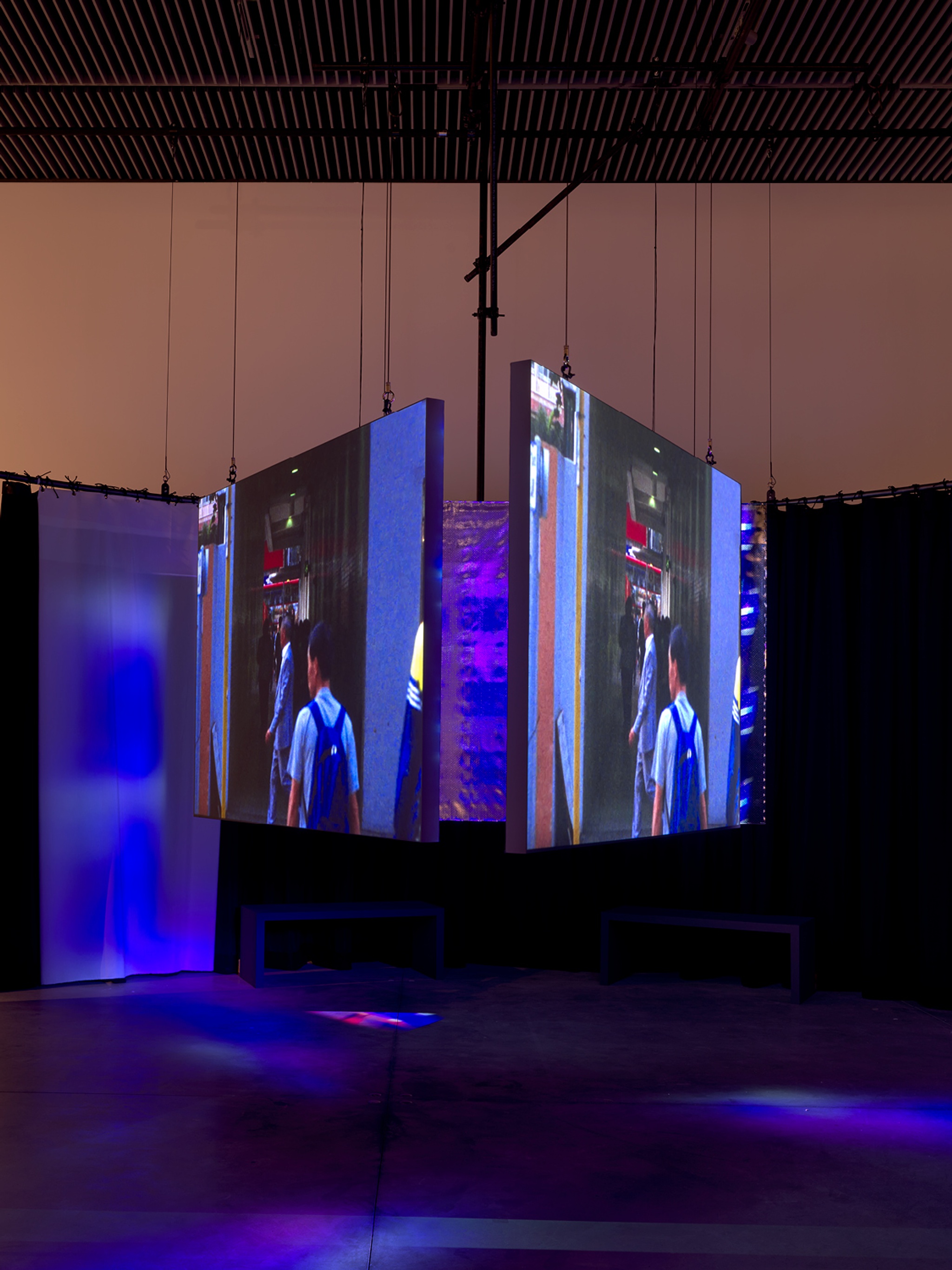 Three video screens hung in a triangular formation in a darkened space awash in bluish light. The screens depict people moving through a cityscape.