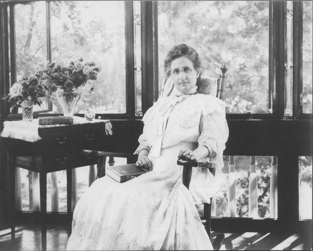 A black and white photograph depicts a light-skinned middle-aged woman a long white dress sitting in a rocking chair in front of several large sunlit windows.