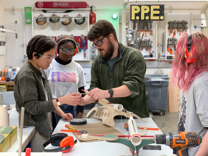 Instructor in a woodshop demonstrates stringing a ping-pong ball crossbow made of wood and PVC to a couple students.