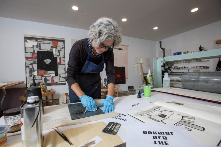 Lisa Bulawsky working in her studio. Numerous papers and drawings are scattered on her table; she is wearing blue gloves while working on a transfer plate. Artworks are hung on the back wall.