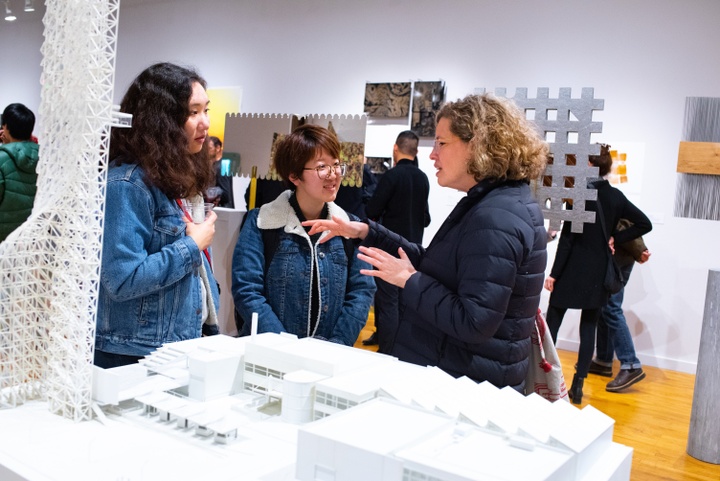 Two students in conversation with director Heather Woofter in the middle of a gallery space. A large white architectural model is in front of them; behind them is additional architectural work and people at the exhibition.