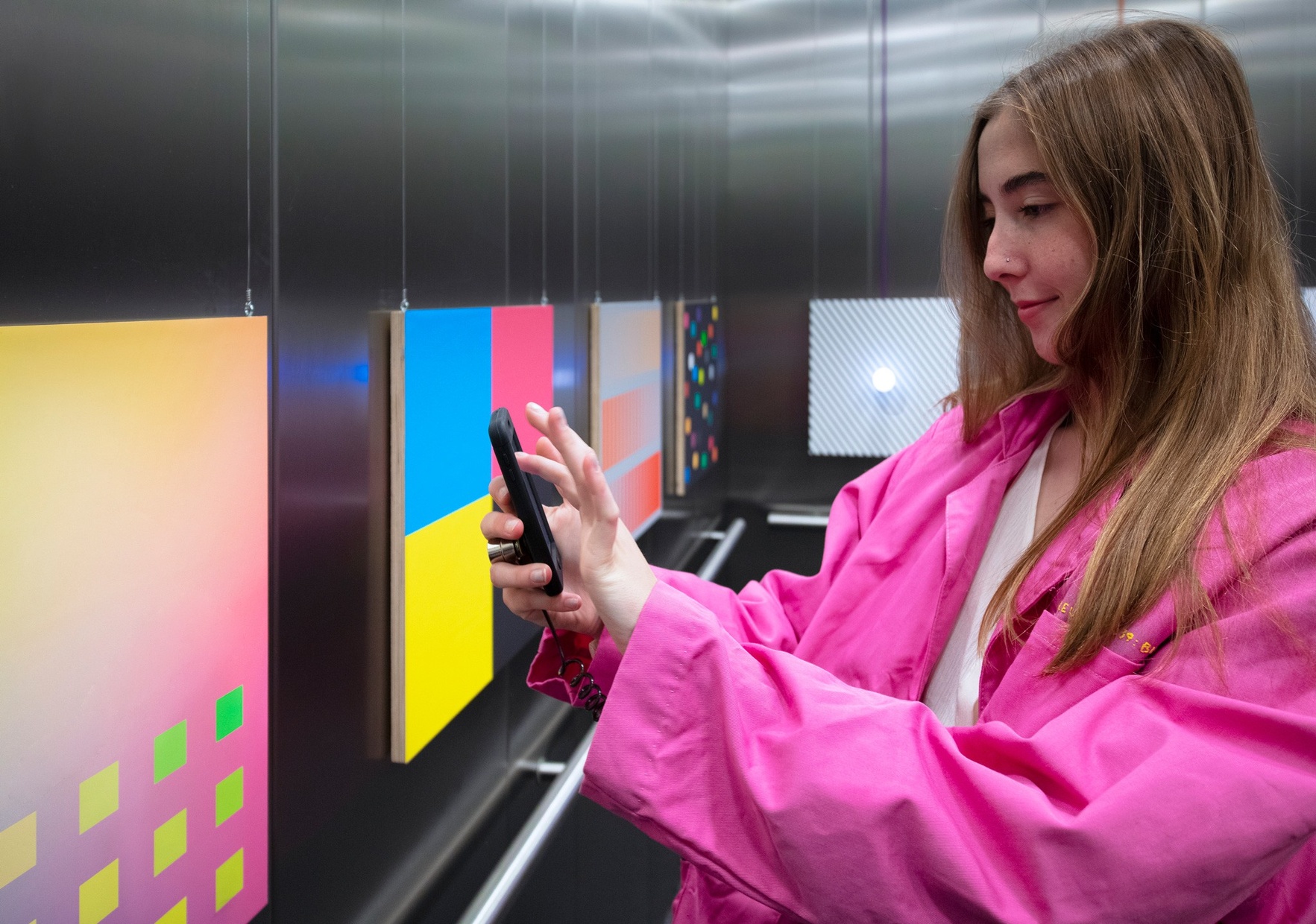 A young, light-skinned female in a bright pink coat is pointing an iphone at a colorful square in an elevator.