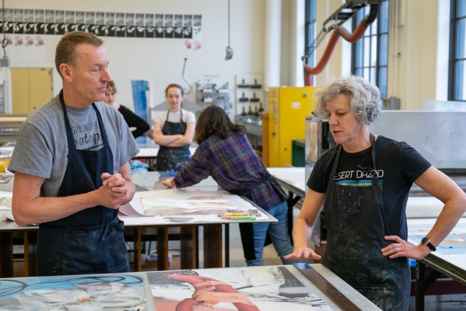 Two people discuss an image collage on a worktable in a printmaking studio while others prepare work behind them.