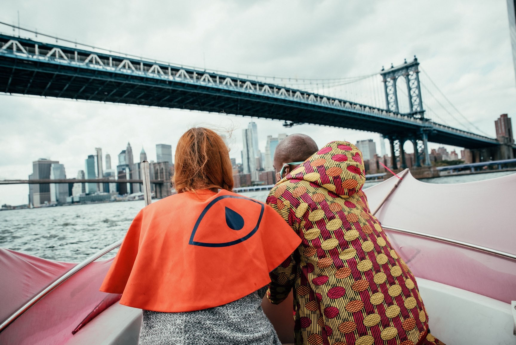 A photograph of two people, backs facing the camera, on a boat overlooking the Brooklyn Bridge. The person on the left wears an orange cape with the Wide Awakes eye.