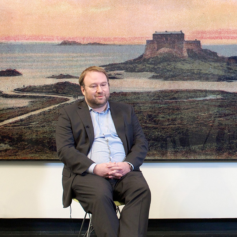 Elger Esser, a white male artist, sits looking down in front of his drawing depicting a sunset behind a large building on an island.