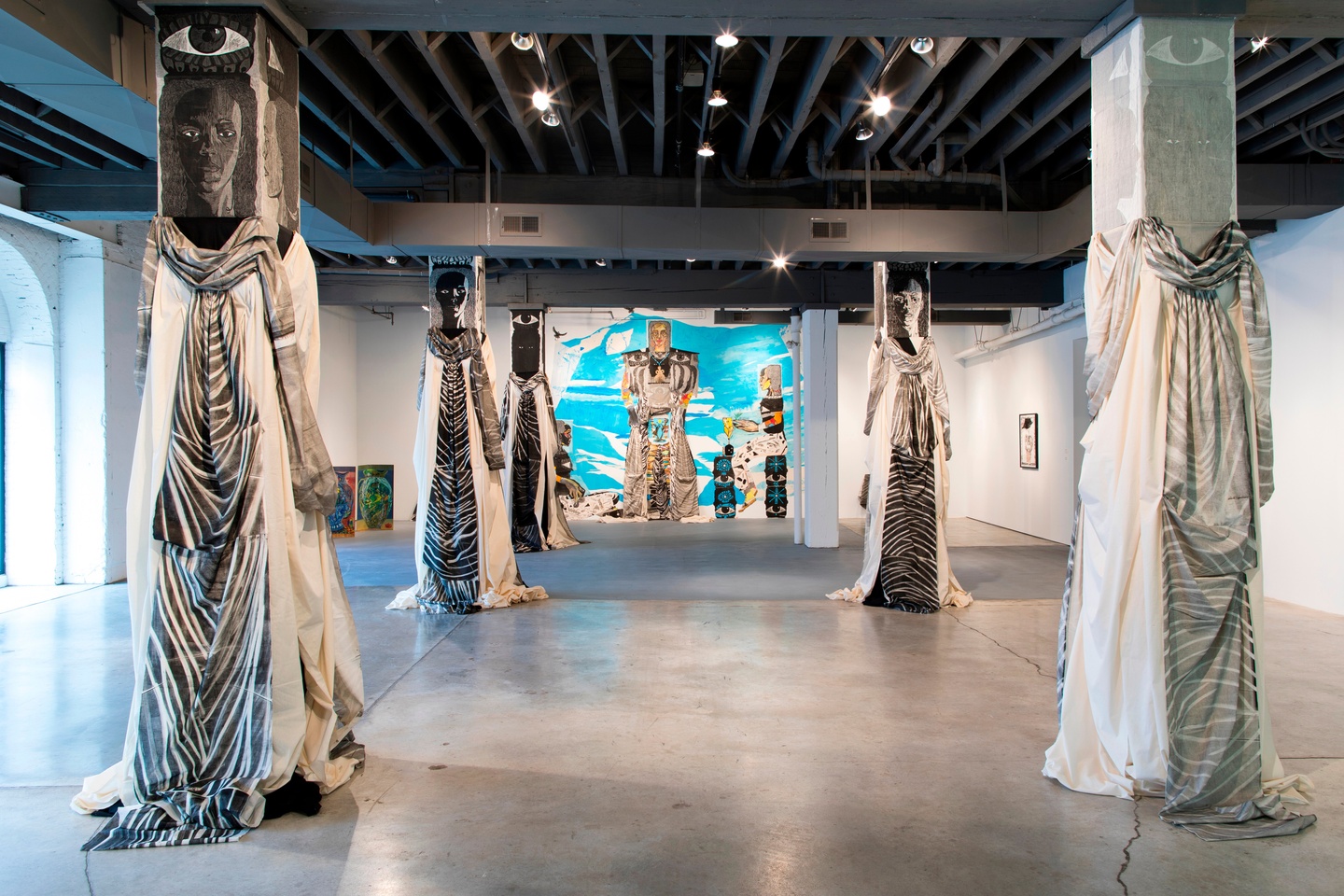 Gallery installation view of works by Paula Wilson, including several pillars draped with fabric, with painted heads to resemble figures, and a painted mural of figures on the back wall.