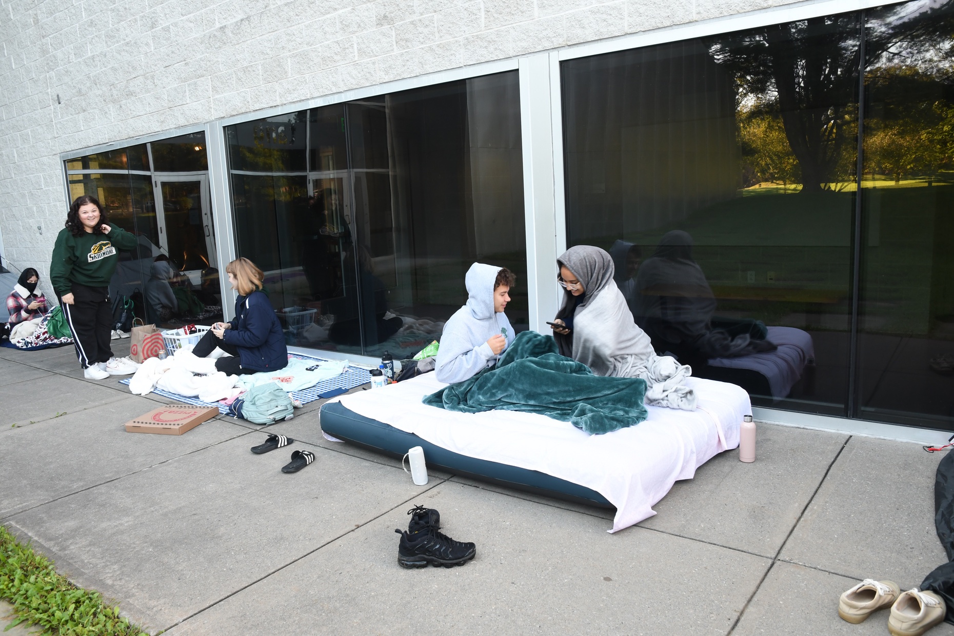A line of students waiting outside the Tang where two students sit on an air mattress with blankets wrapped around them.