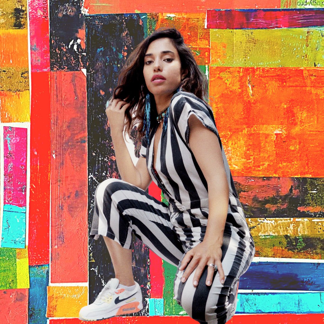 A portrait of DJ L3NI kneeling in front of a colorful wall with a rectangular color block design. She wears a jumpsuit with black and white vertical stripes.