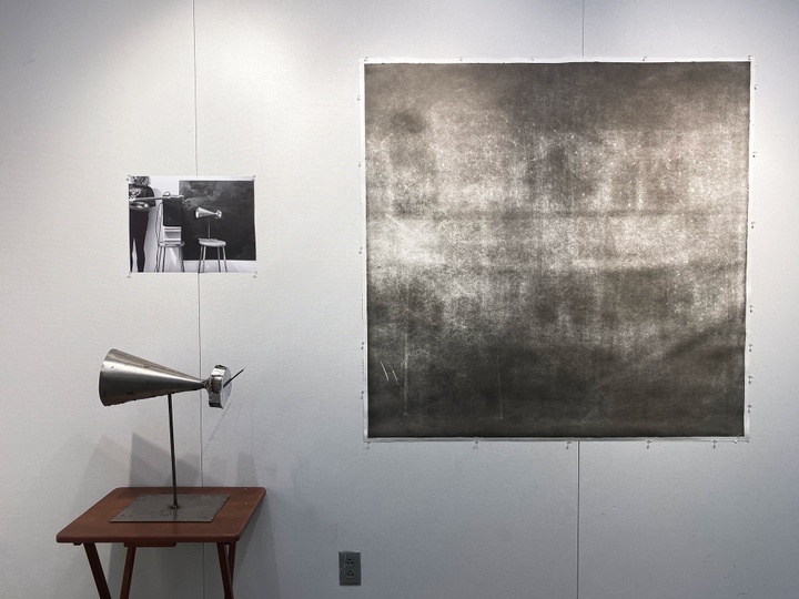 a metal megaphone on a stand on a table. The wall behind it has a photograph of a similar scene and a larger print of black on newsprint