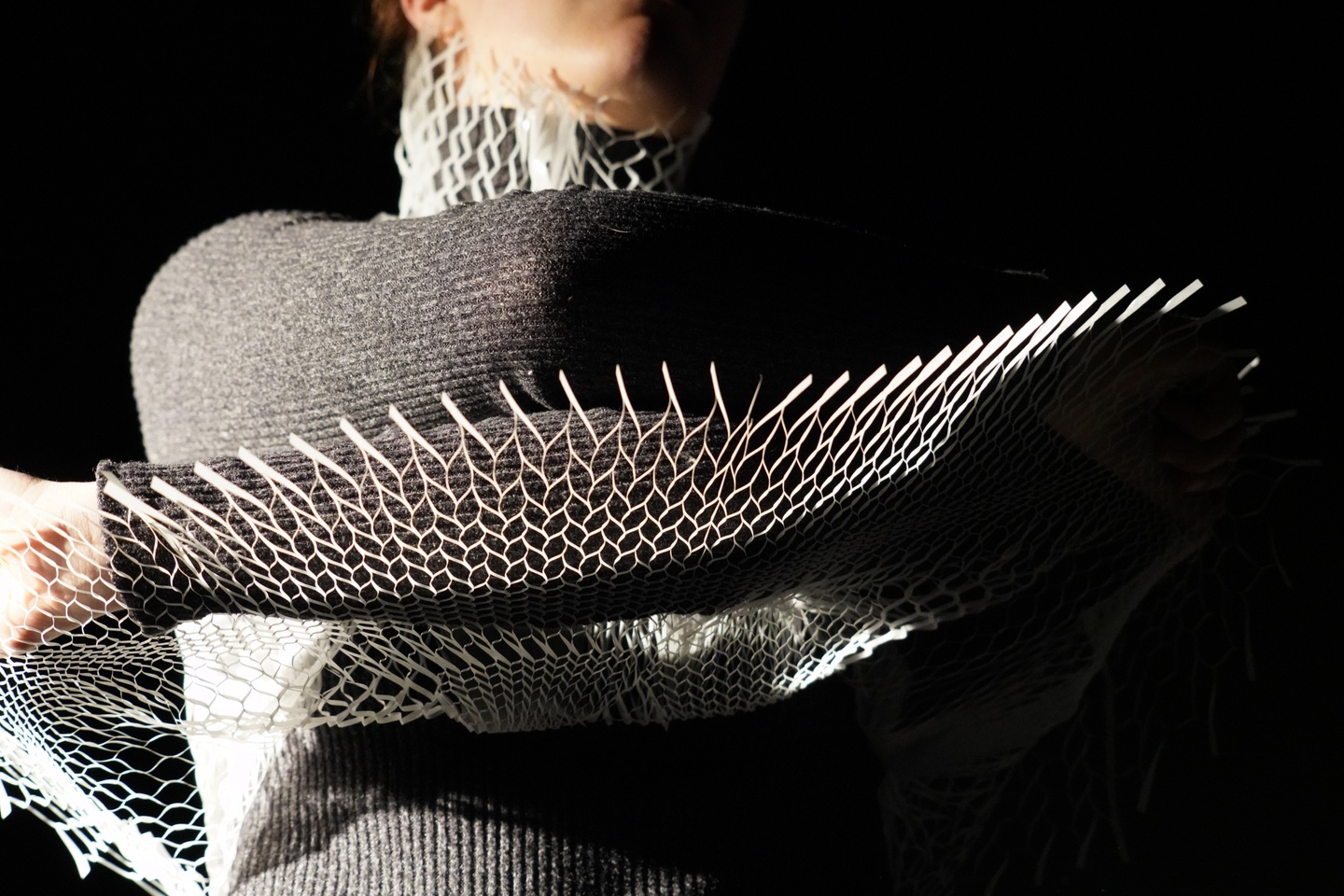 View of a person's torso wearing a grey turtleneck grabbing the bottom edges of a white 3D printed mesh shirt and lifting it up as if to take it off.