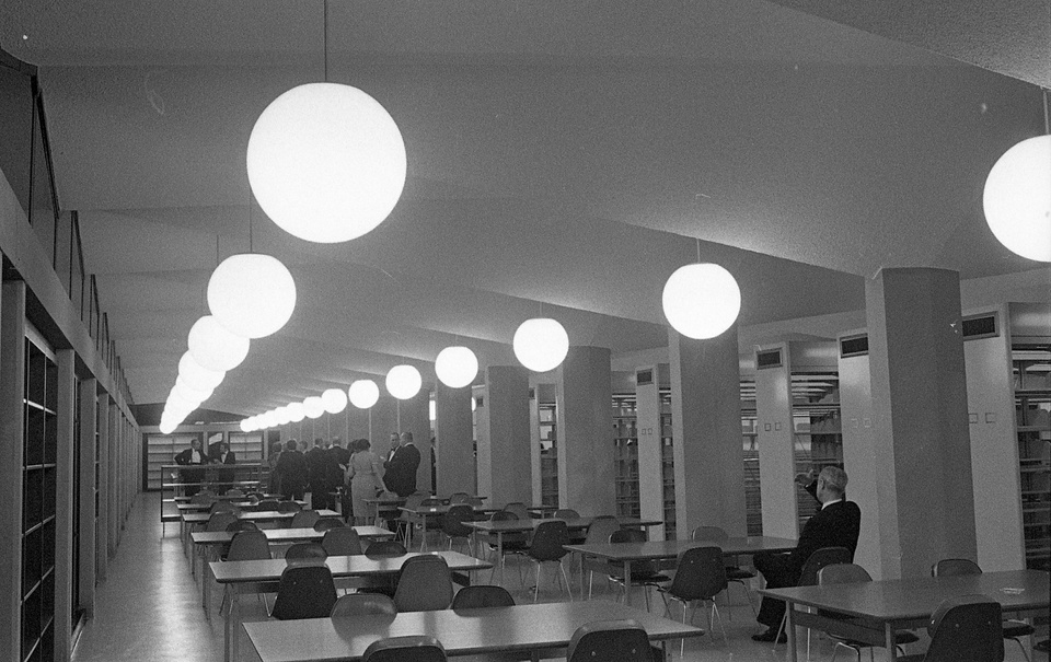 Black and white photo of a long room with a pleated ceiling and huge globe lights. There are rows of tables and chairs, and library stacks line the left wall.