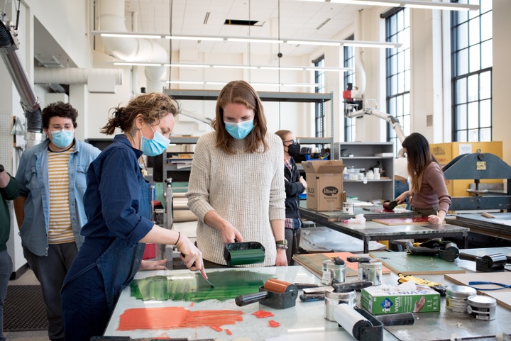 Large printmaking studio space. Two people stand at a table. One uses a putty knife to remove green ink from a glass tabletop.