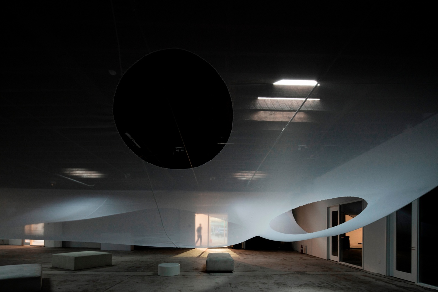 An oversize textile with a couple of large holes cut out hangs from the ceiling of an open industrial space with a few pieces of minimalist furniture on the ground.