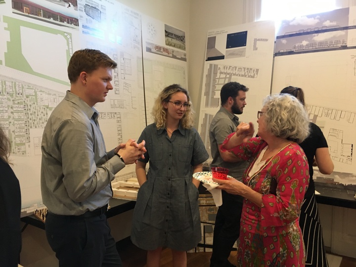 Three people stand in conversation in front of boards of architectural drawings. 