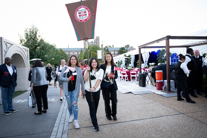 Three smiling students walk down a pathway carrying a brown banner with the WashU seal on it.