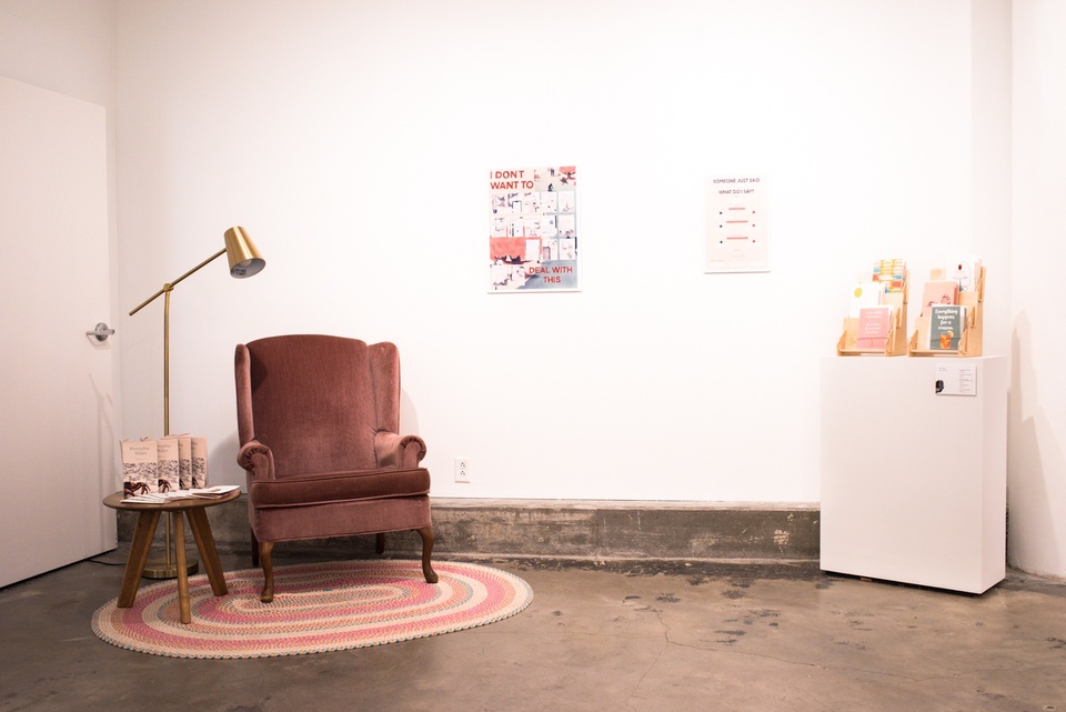Installation with a velour armchair, braid rug, end table, lamp, and brochure rack. A poster on the wall reads "I don't want to deal with this" and contains comics panels.